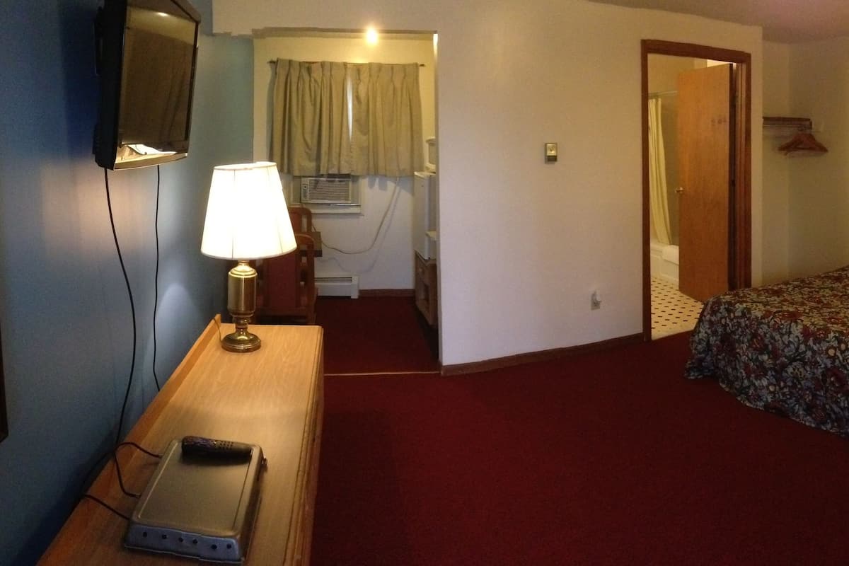 A hotel room with a bed, a desk with a lamp, a chair, a small table, and a television.