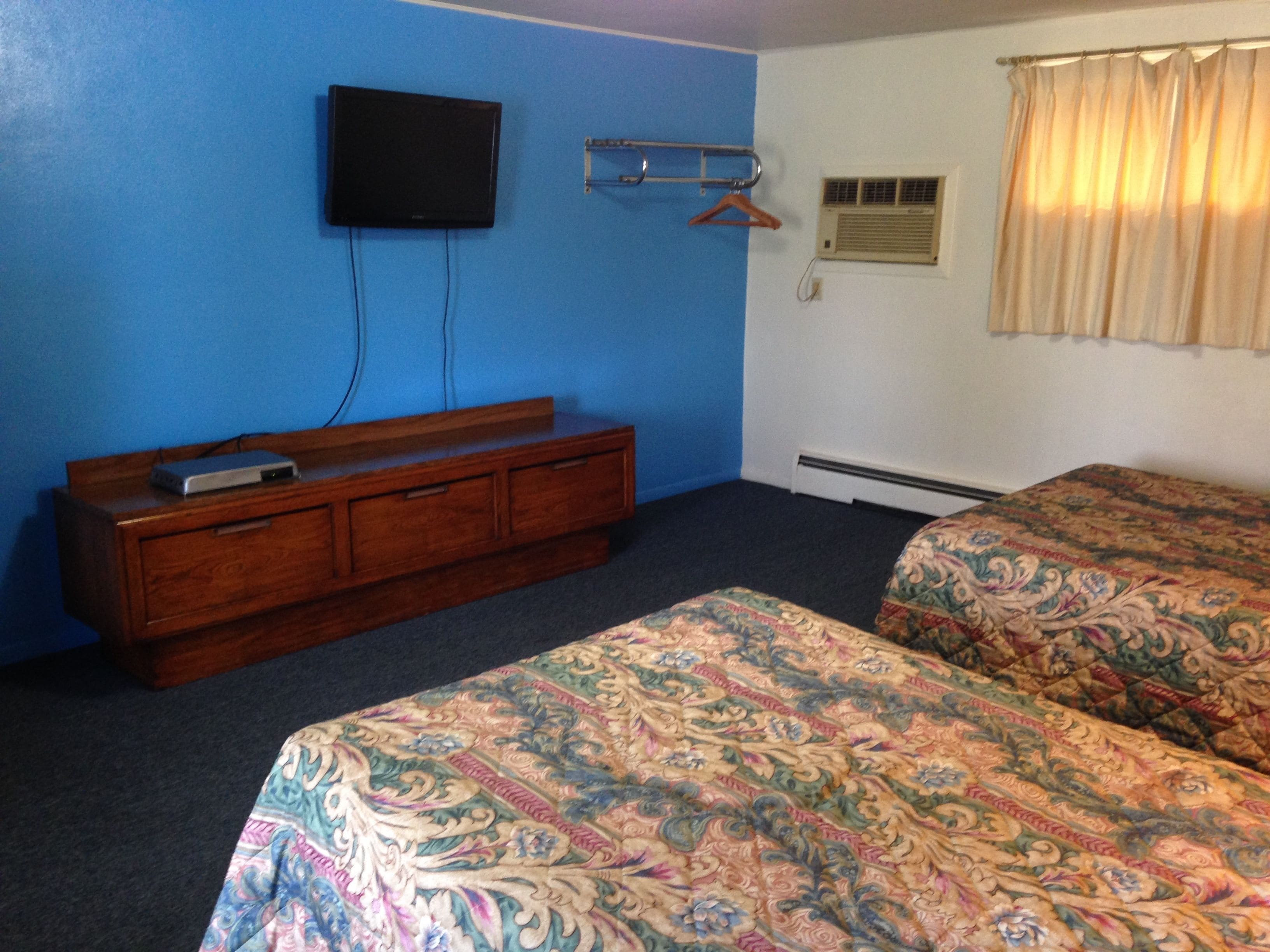 Habitación de hotel con dos camas, un televisor montado en la pared, un aparador de madera y una ventana con cortinas.