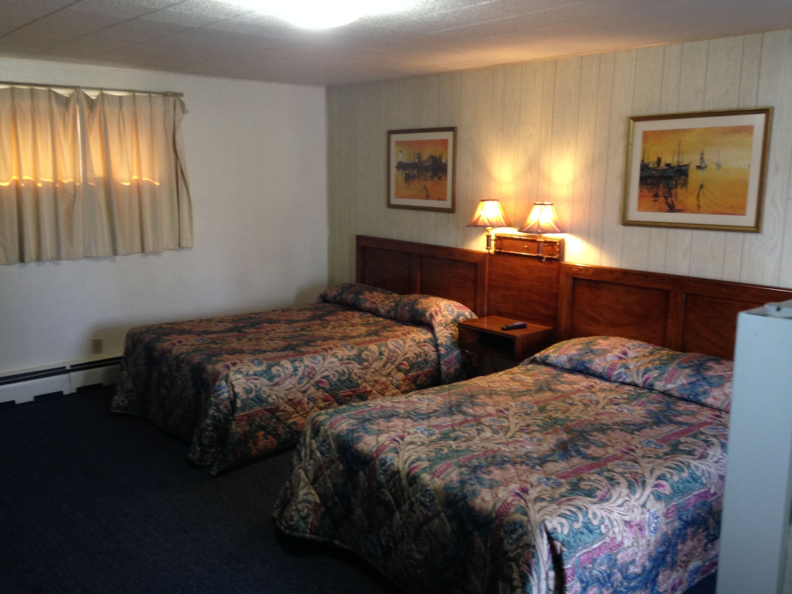 Habitación de hotel con dos camas, una mesita de noche, dos lámparas fijadas a la pared y dos cuadros enmarcados.