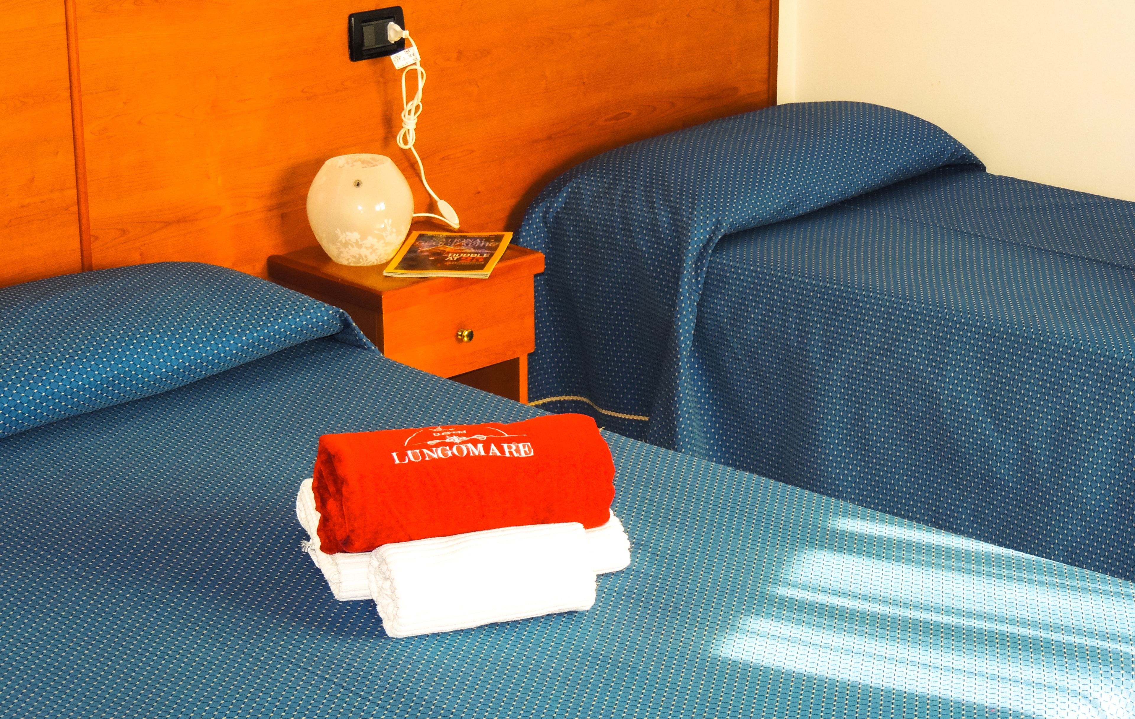 Camera d'albergo con testiera in legno, un comodino con lampada, un libro e un asciugamano piegato rosso e bianco sul letto.