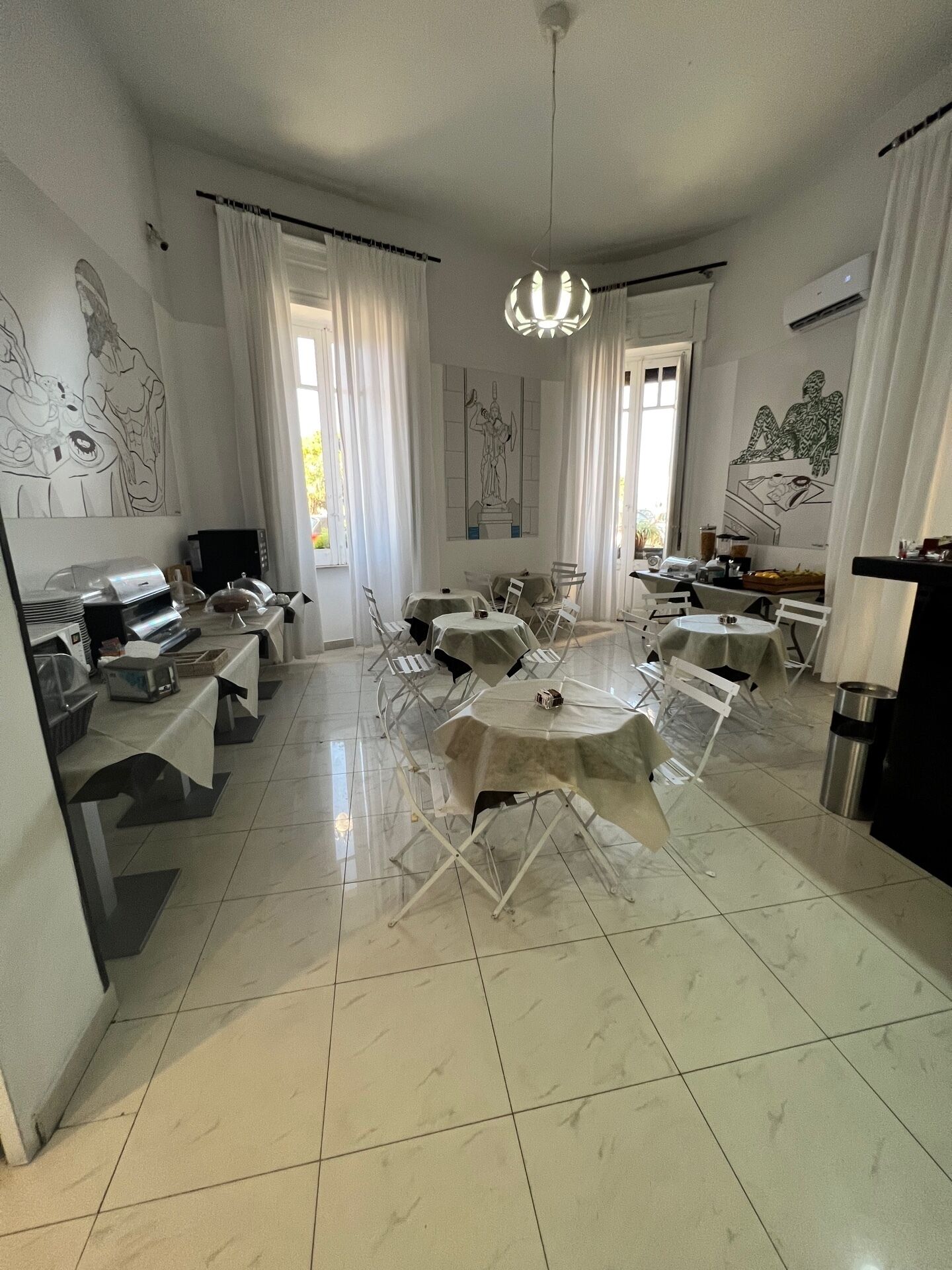 A dining area with a round table, chairs, and a chandelier. There are white curtains, a tiled floor, and artwork on the walls.