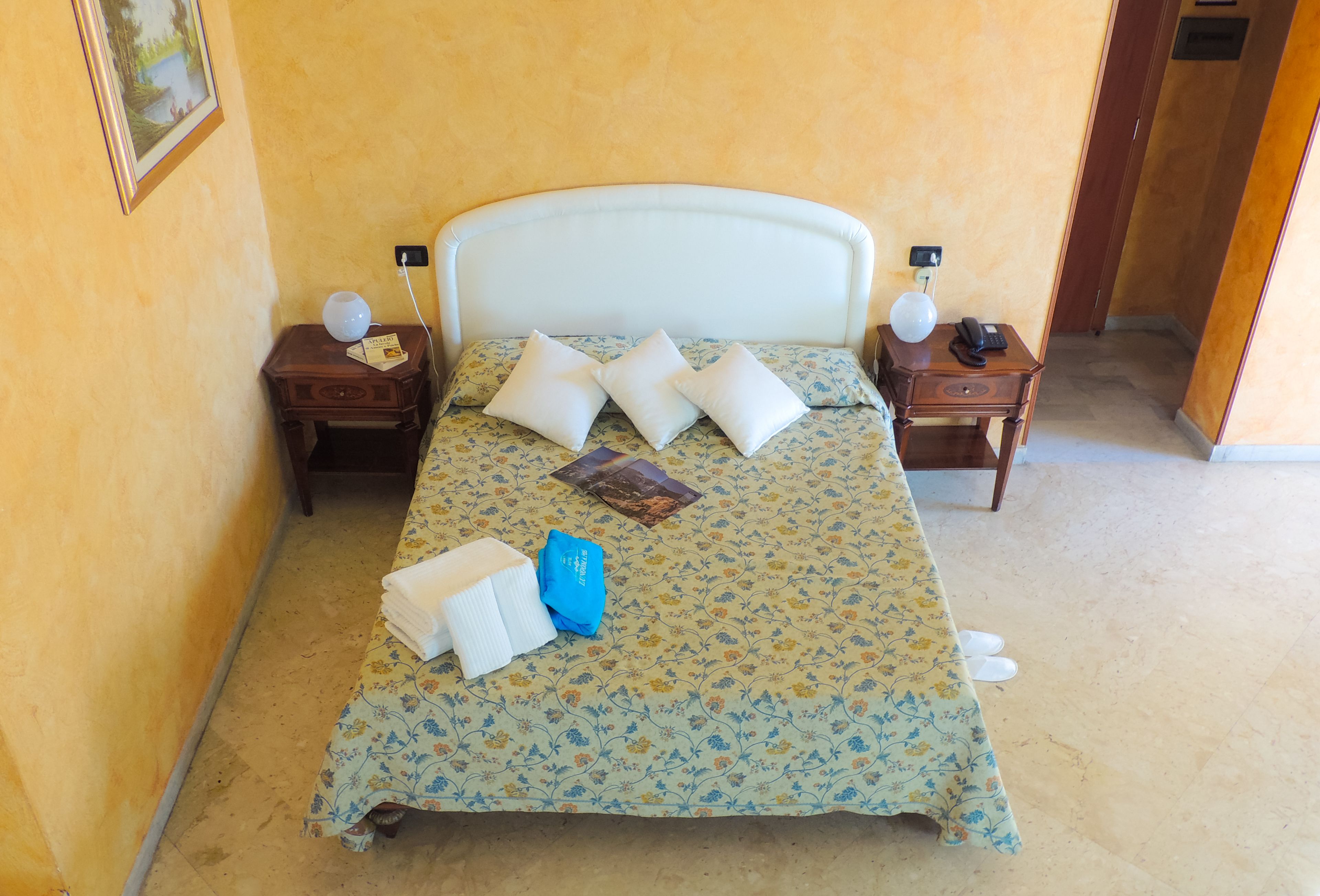 Una cama bien hecha con una colcha floral, dos almohadas, una toalla doblada y una prenda azul sobre la cama. Hay dos mesitas de noche con lámparas y un teléfono.