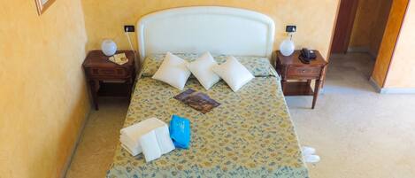 Una cama bien hecha con una colcha floral, dos almohadas, una toalla doblada y una prenda azul sobre la cama. Hay dos mesitas de noche con lámparas y un teléfono.