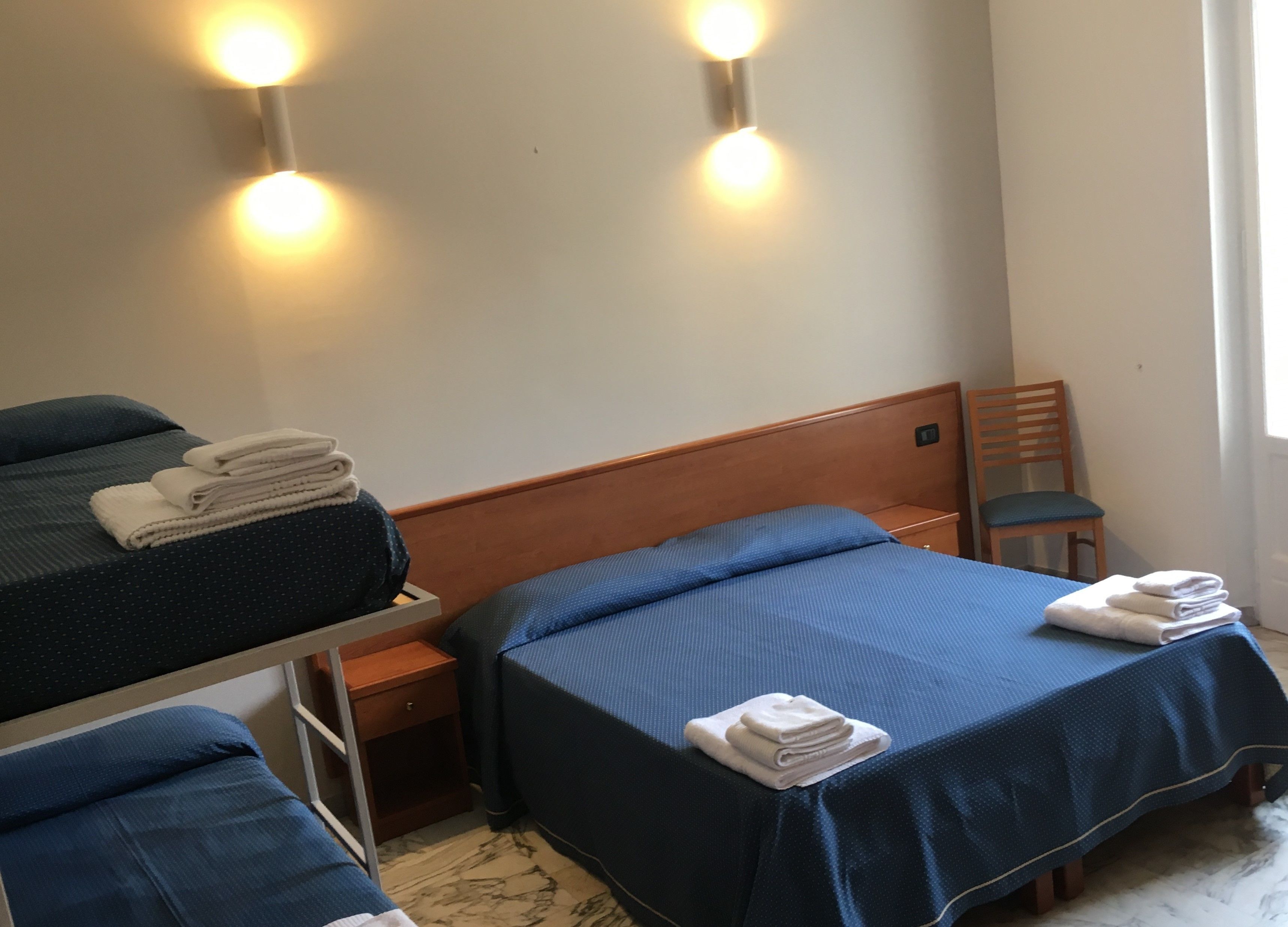 Habitación de hotel con dos camas, cada una con colcha azul y toallas blancas. Hay luces fijadas a la pared sobre las camas, una mesita de noche de madera y una silla.