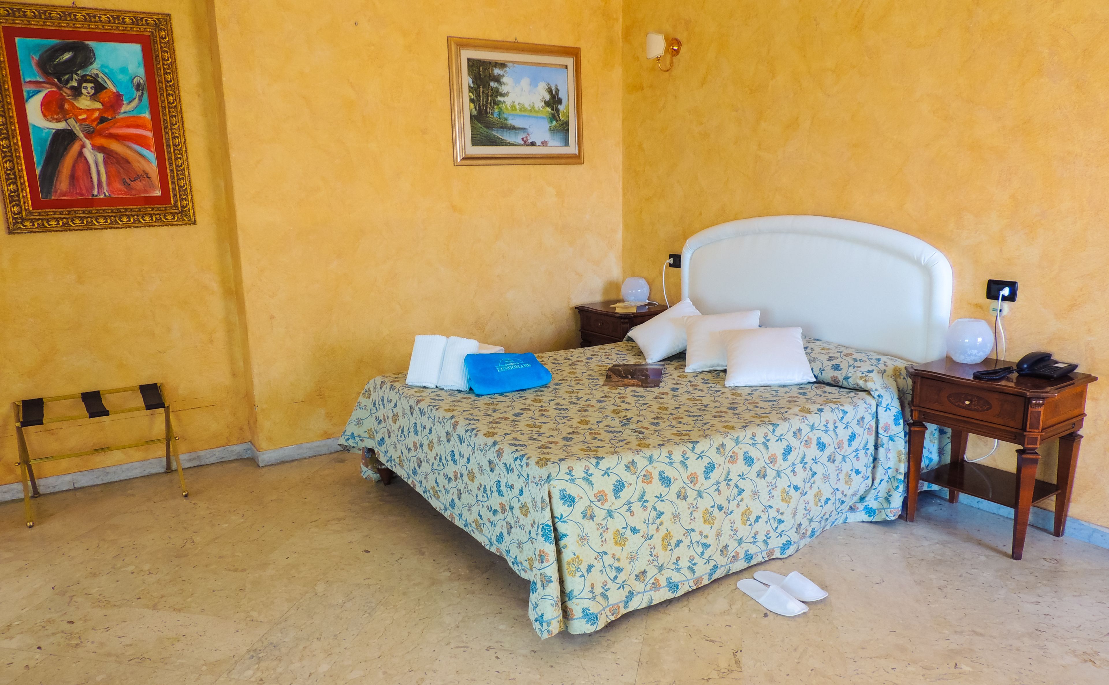 A bedroom with a bed, a nightstand, a lamp, a painting, and a framed picture on the wall.