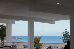 Una terraza con columnas blancas, mobiliario exterior y vistas al mar y montañas lejanas.