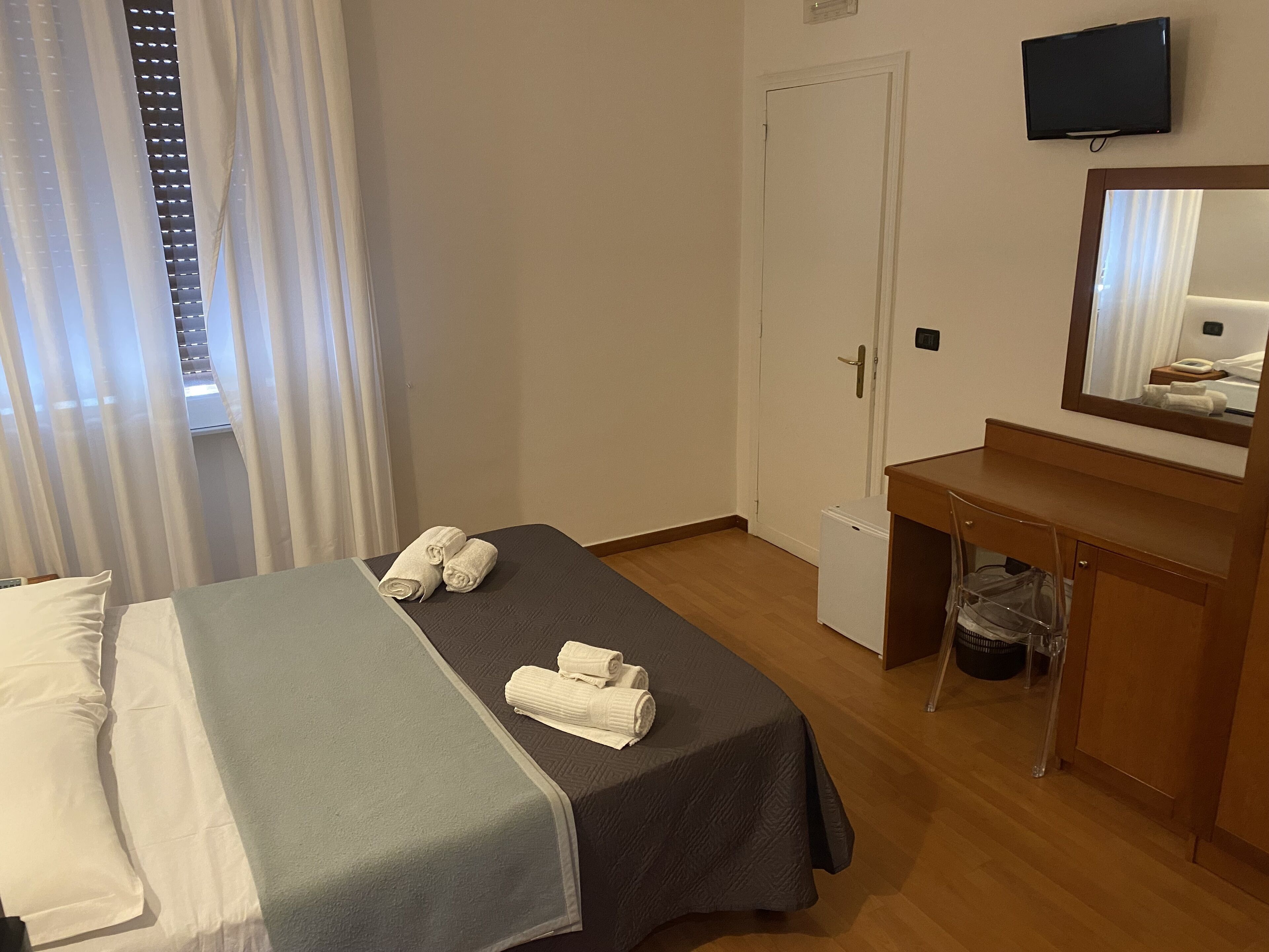 A hotel room with a bed, a small desk, a mirror, a television, and a window with curtains.