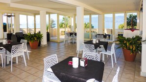 A spacious indoor dining area with multiple tables and chairs, large windows offering a view of the sea, and potted plants. - Lungomare Hotel (Reggio Calabria)