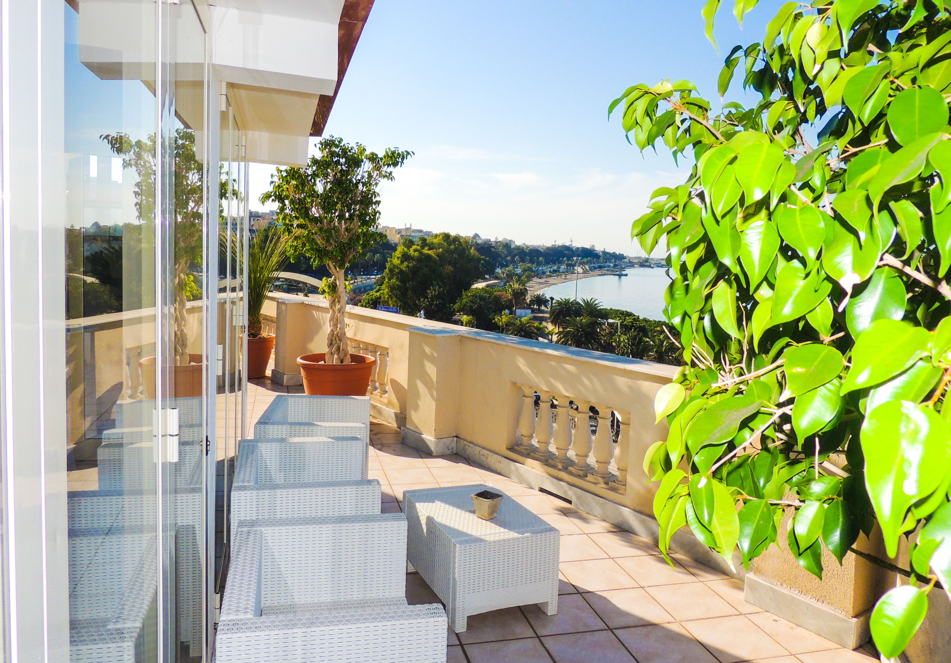 A balcony with white wicker furniture, potted plants, and a view of a body of water and distant buildings.