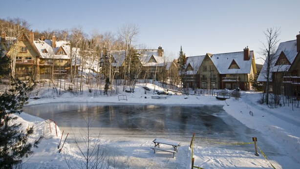 A snow-covered outdoor area with a frozen pond, benches, and a picnic table. - Le Plateau – RVMT (Mont-Tremblant)