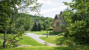 A house with a driveway leading to a front door, surrounded by lush greenery and trees. - Le Plateau – RVMT (Mont-Tremblant)