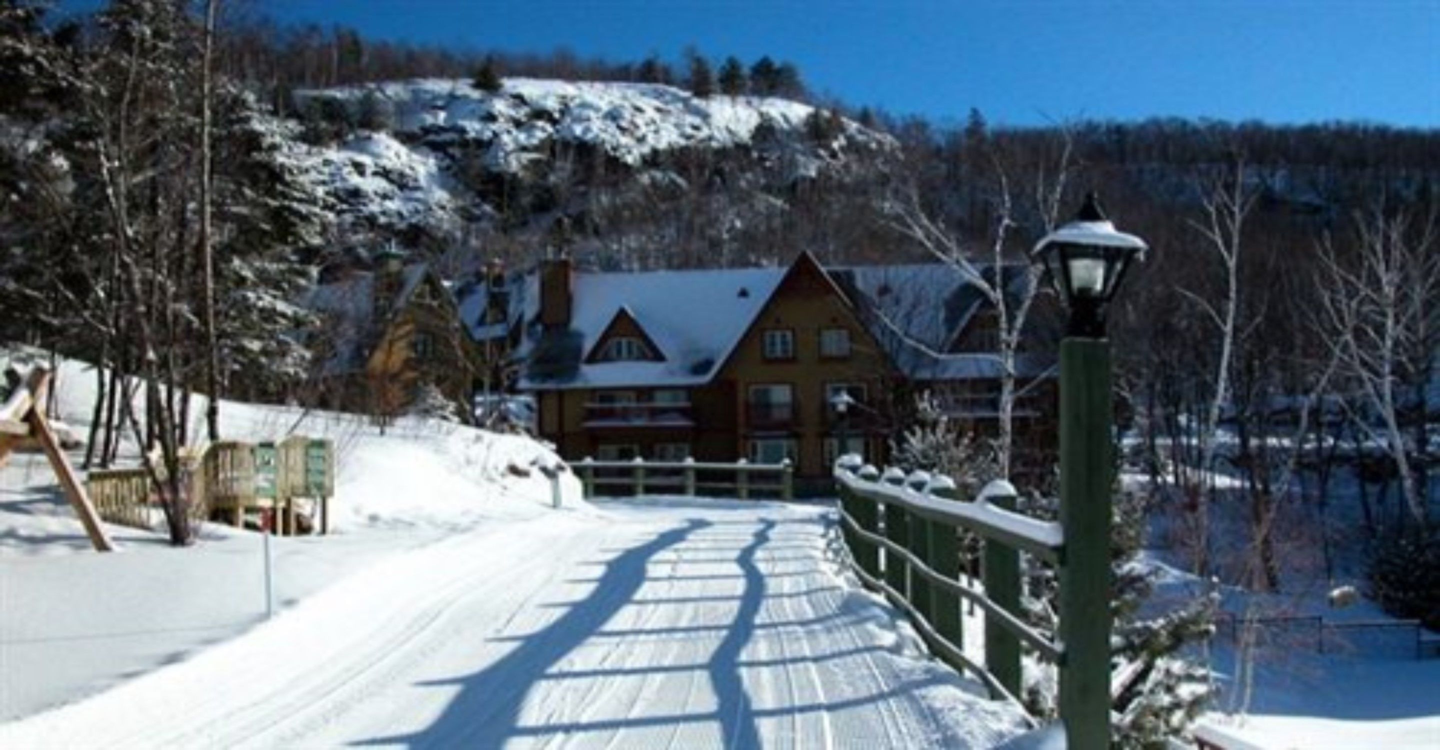 A snow-covered pathway leading to a wooden lodge with a snow-covered roof, surrounded by trees and a hill in the background.