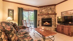 A living room with a stone fireplace, a sofa, a table, and a television. - Le Plateau – RVMT (Mont-Tremblant)