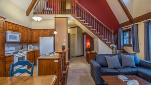 A spacious kitchen with wooden cabinets, a microwave, and a refrigerator. A wooden dining table and chairs are positioned near the kitchen. A staircase leads to a loft area with a red ceiling. - Le Plateau – RVMT (Mont-Tremblant)