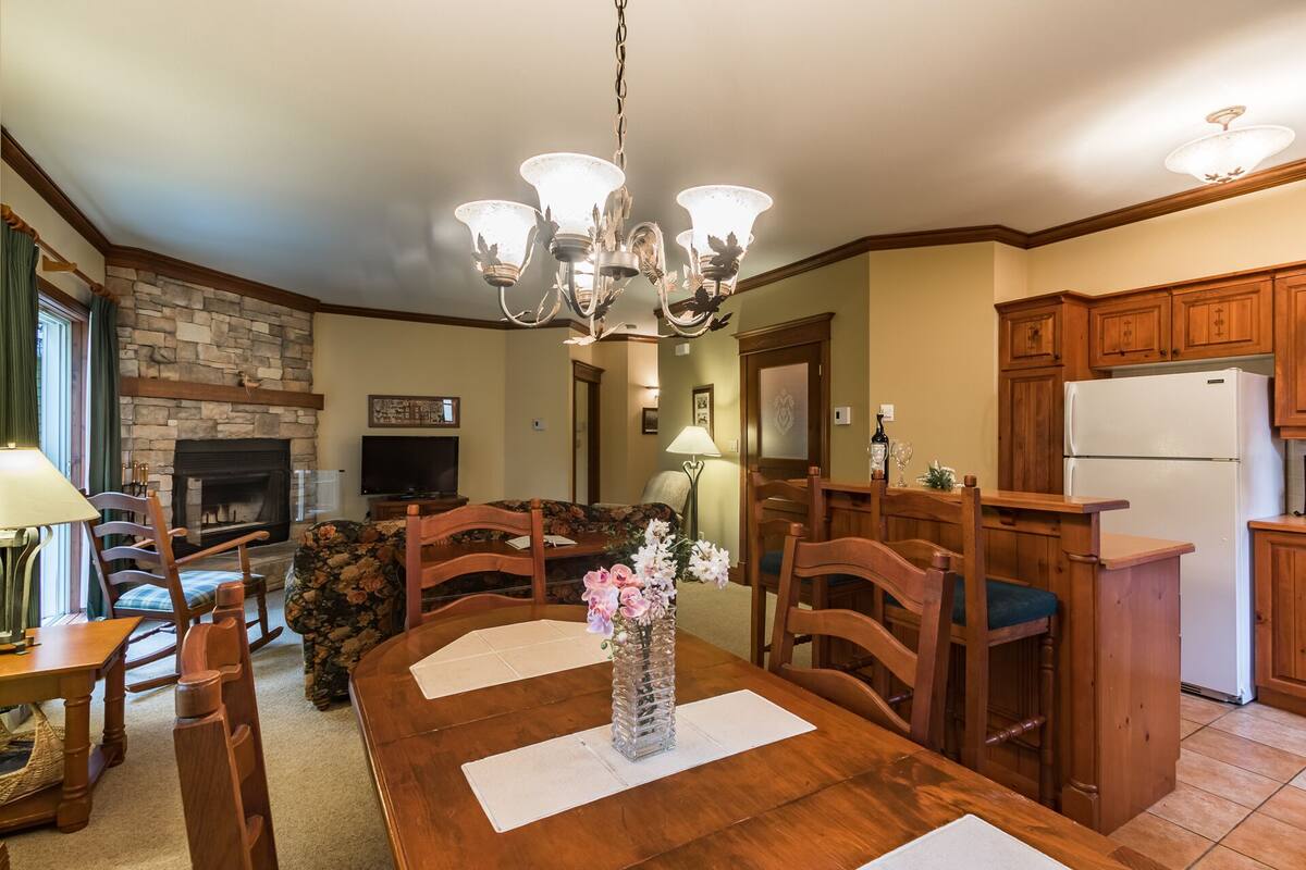 Un comedor con una mesa de madera, sillas, un candelabro, una chimenea y una cocina con armarios de madera y un refrigerador.