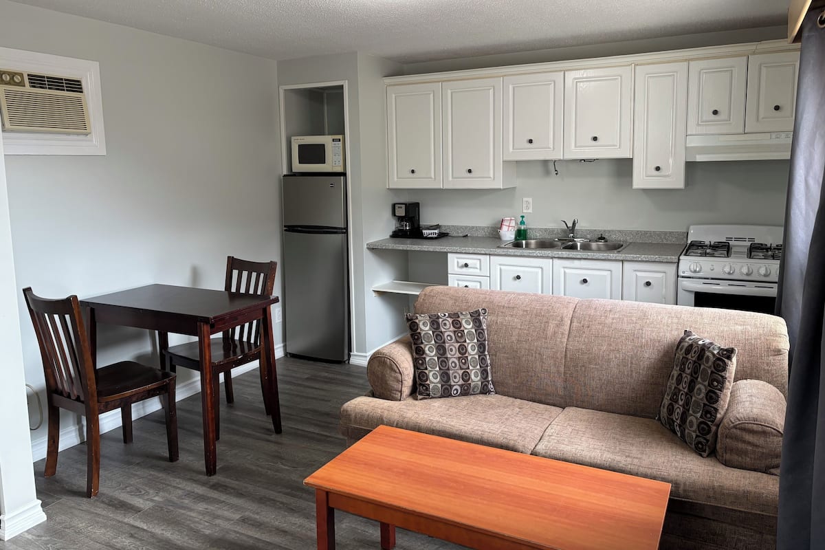 A compact kitchen with white cabinets, a microwave, a refrigerator, and a stove. There is a dining area with a wooden table and chairs, and a sofa with patterned cushions.