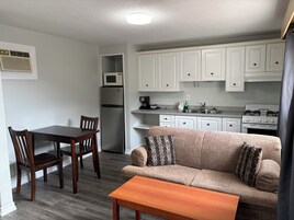 Cocina compacta con armarios blancos, microondas, refrigerador y estufa. Hay un comedor con mesa y sillas de madera, y un sofá con cojines estampados.