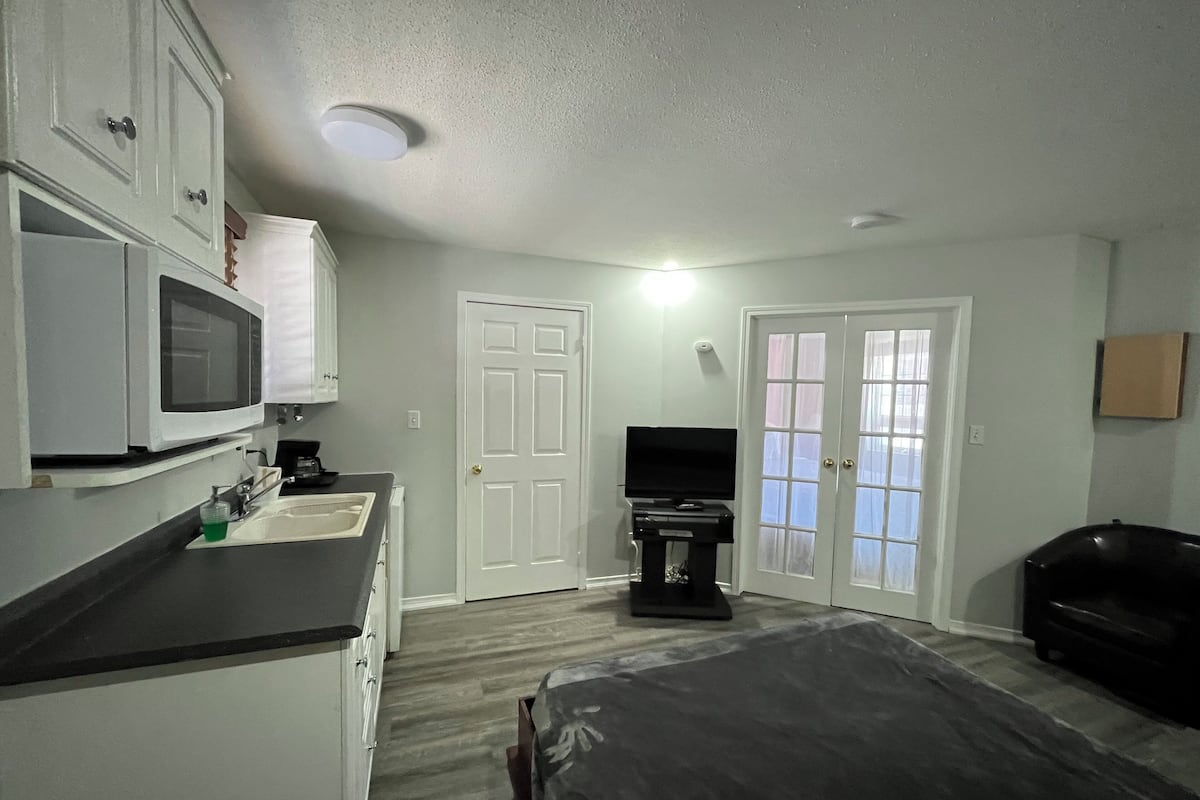 A compact room with a kitchenette, a bed, a TV, and a door with frosted glass panels.