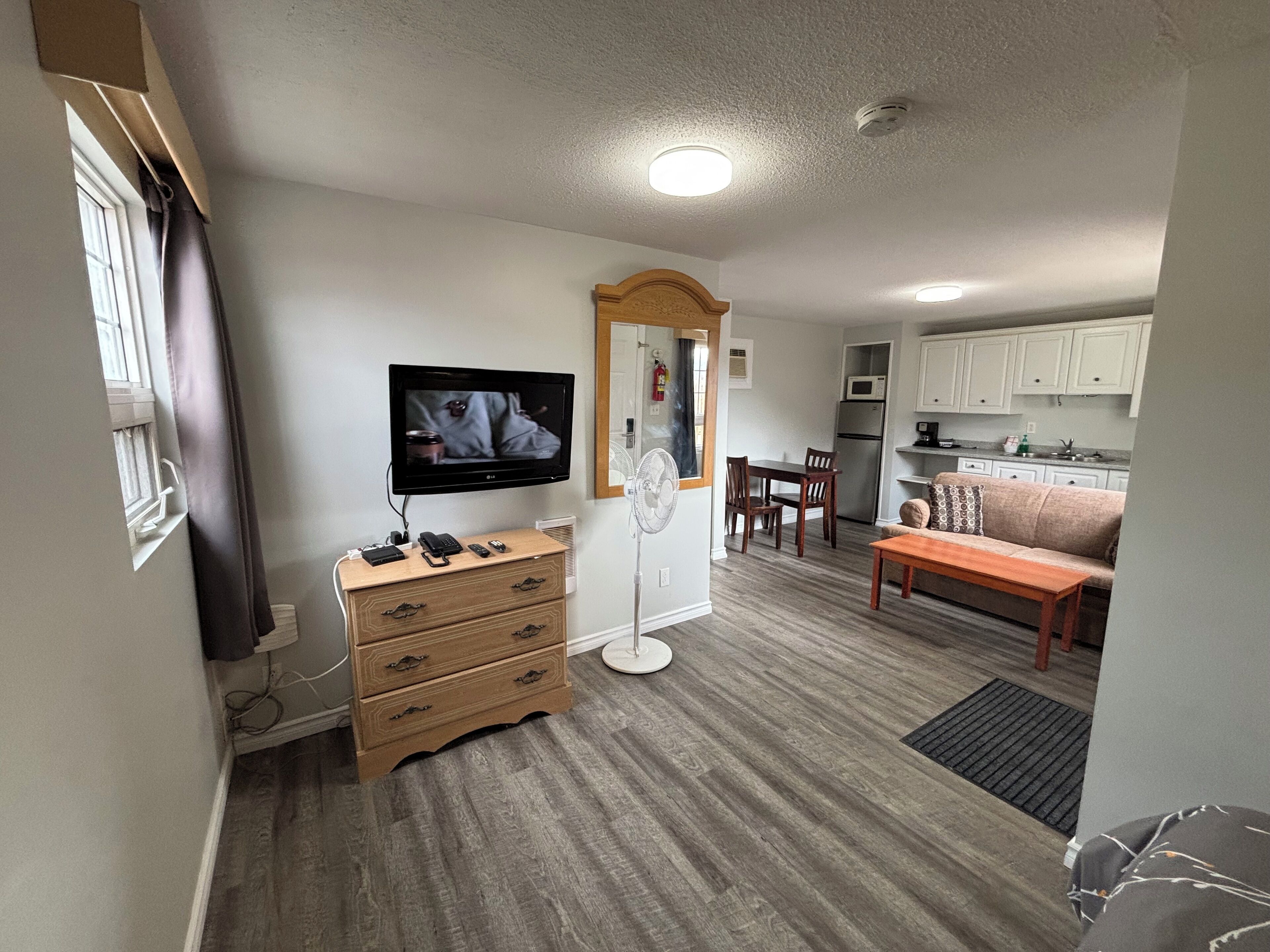 Un espace de vie compact comprenant un téléviseur à écran plat fixé au mur, une commode en bois, une table à manger avec des chaises, un canapé et un coin cuisine.