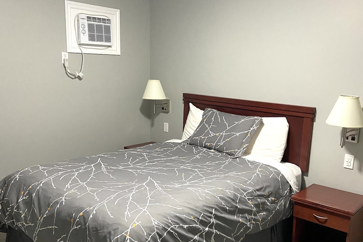 A neatly made bed with a gray and white patterned bedspread, two bedside lamps, a wooden nightstand, and a wall-mounted air conditioning unit.