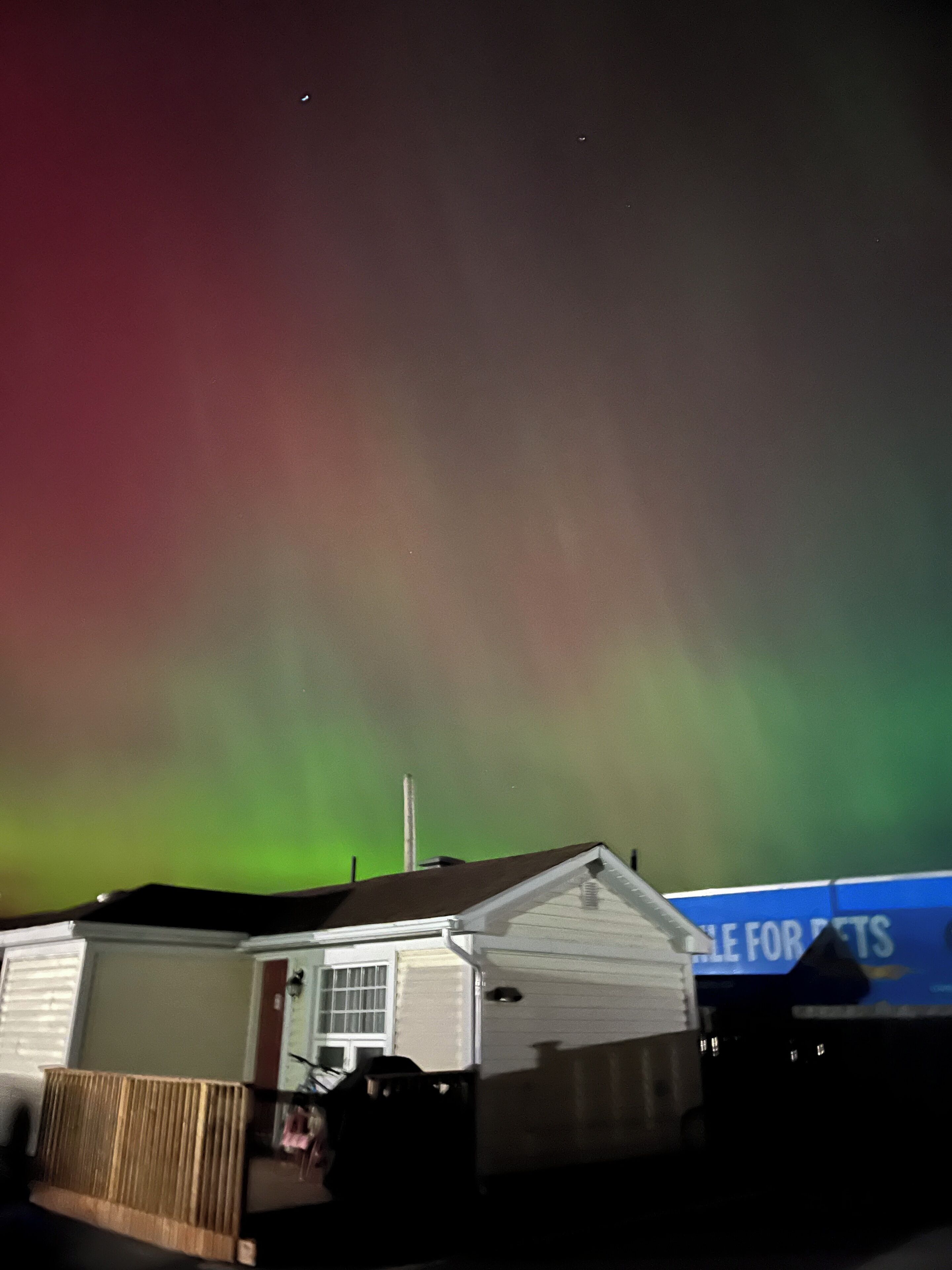 Une maison avec une cheminée bien visible, une cour clôturée et un ciel nocturne dégagé illuminé par les aurores boréales.