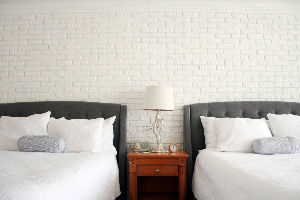 A hotel room with two beds, white bedding, a wooden nightstand with a lamp, a clock, and a power outlet.