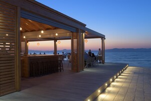 Zona de bar al aire libre con vigas de madera y luces colgantes, con vistas a una masa de agua.