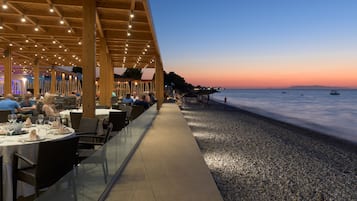 An outdoor dining area with tables and chairs, overlooking a pebble beach and the sea.