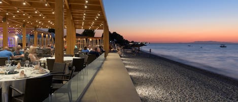 An outdoor dining area with tables and chairs, overlooking a pebble beach and the sea.