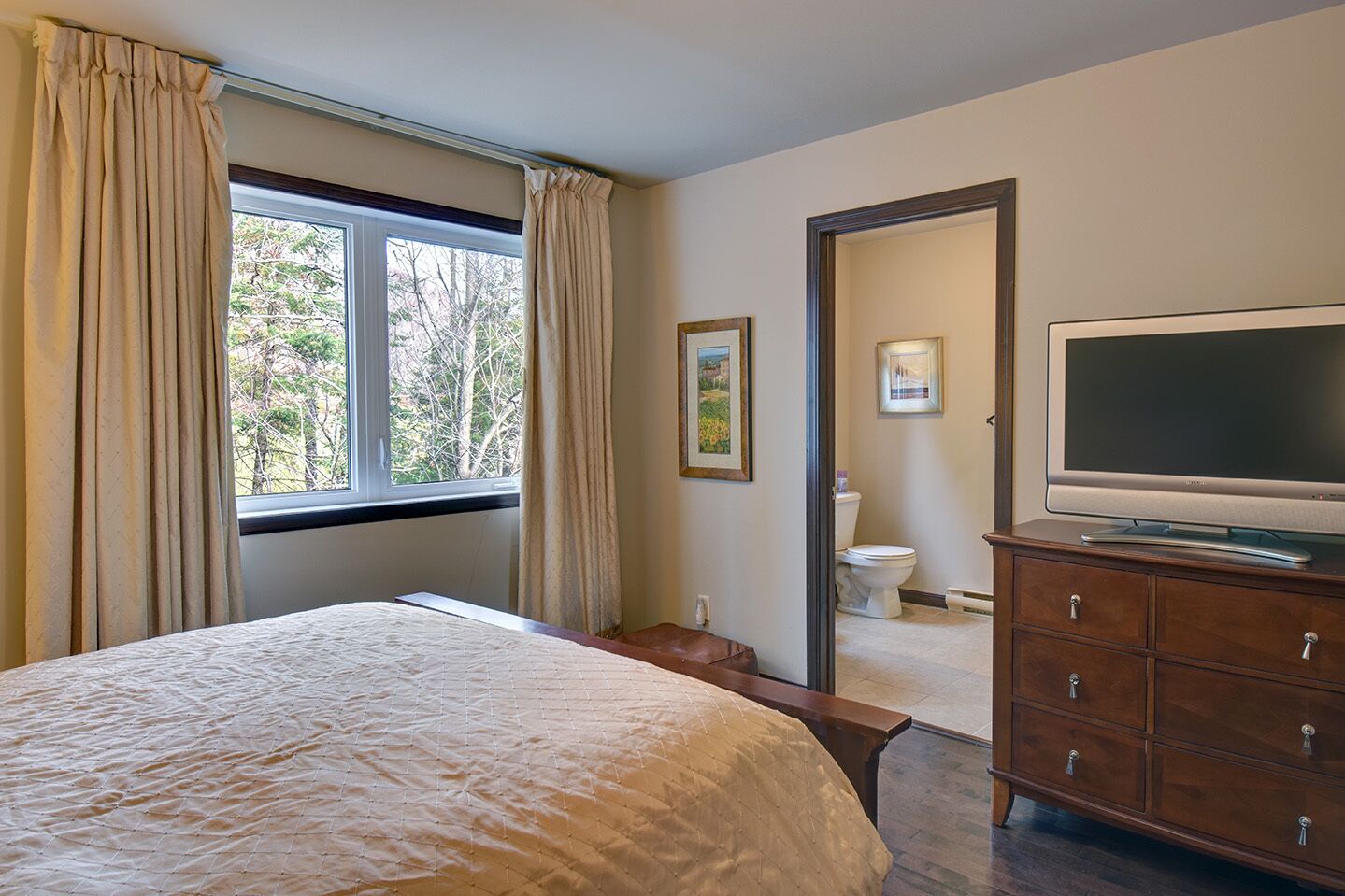 A bedroom with a large bed, a television on a wooden dresser, and a window with curtains.