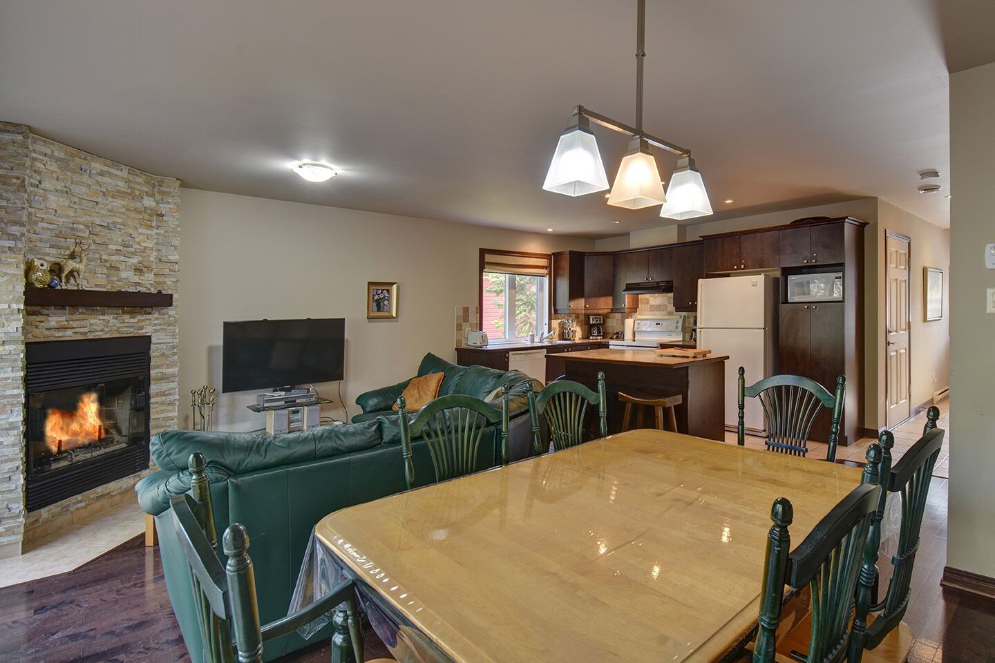 A spacious living room with a fireplace, a green sofa, a wooden dining table, and a kitchen area with dark wood cabinets and a refrigerator.