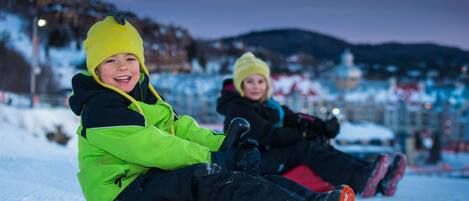 Two children sledding on a snowy slope with a mountainous backdrop.