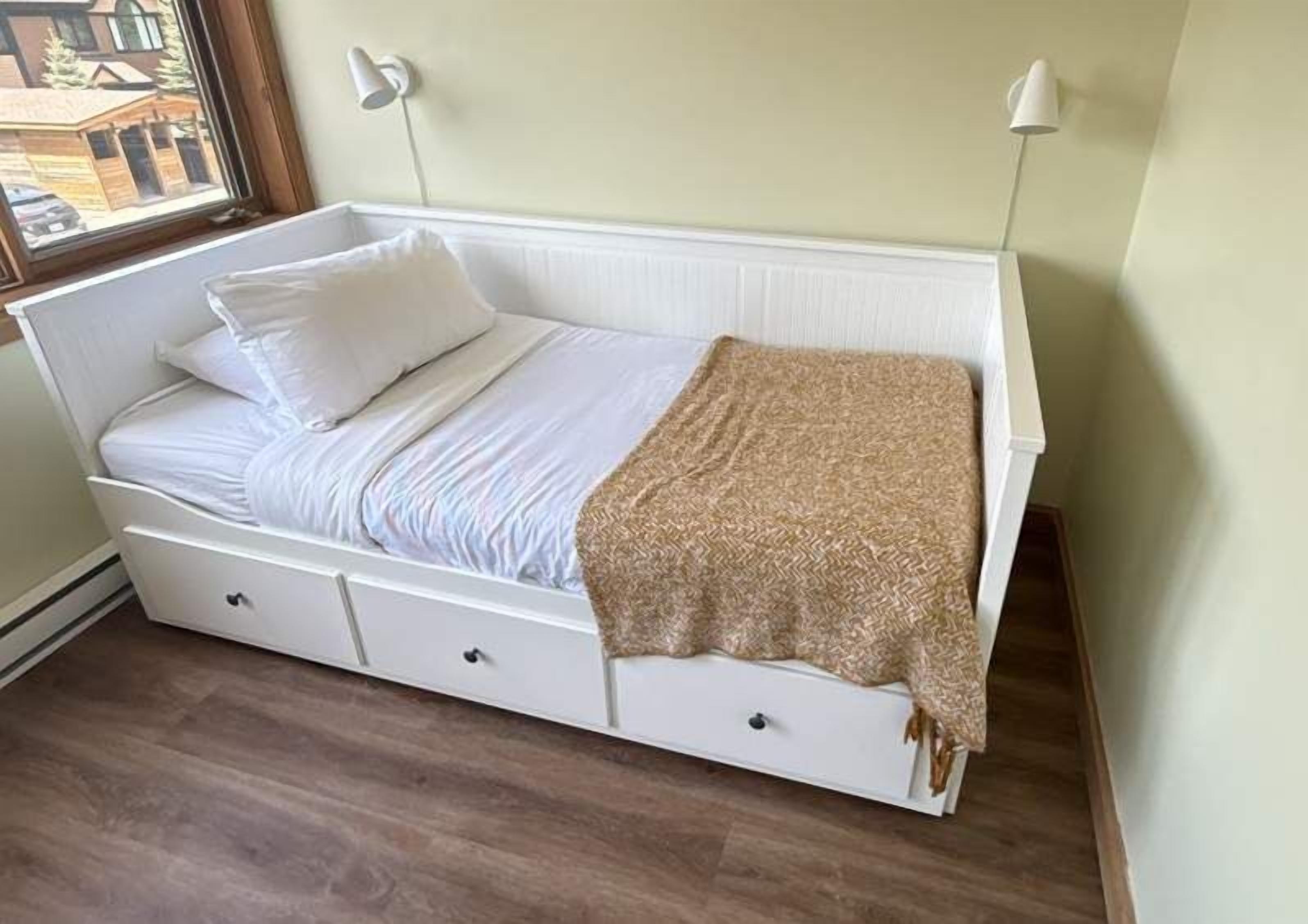 A single-bed room with a white headboard, a bedside table, a drawer, and a window with a view of buildings.