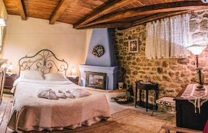 A cozy bedroom with a stone wall, a fireplace, a bed with a headboard, and a nightstand with a lamp.