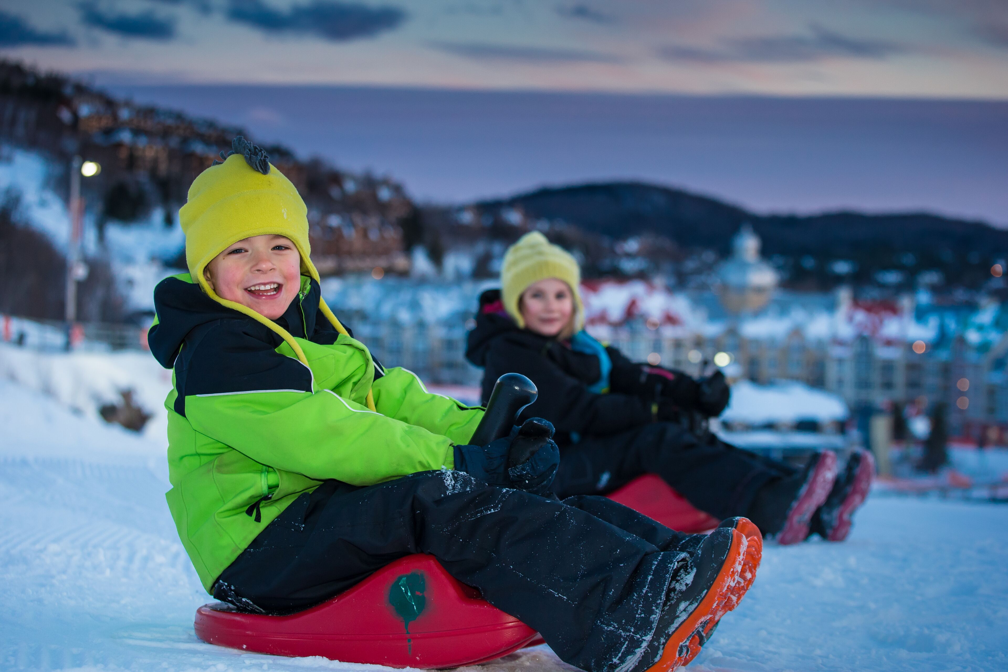 Deux enfants dévalent une pente enneigée en luge, avec une chaîne de montagnes en arrière-plan.