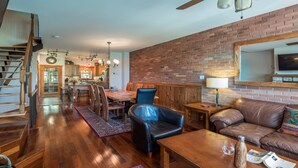 Amplia sala de estar con escalera de madera, una mesa de comedor con sillas, sofá de cuero y una pared de ladrillo decorativa.