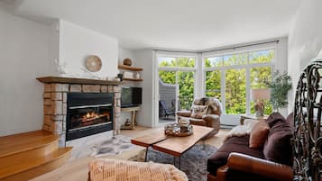 A living room with a stone fireplace, a television, a sofa, and a large window with a view of trees.