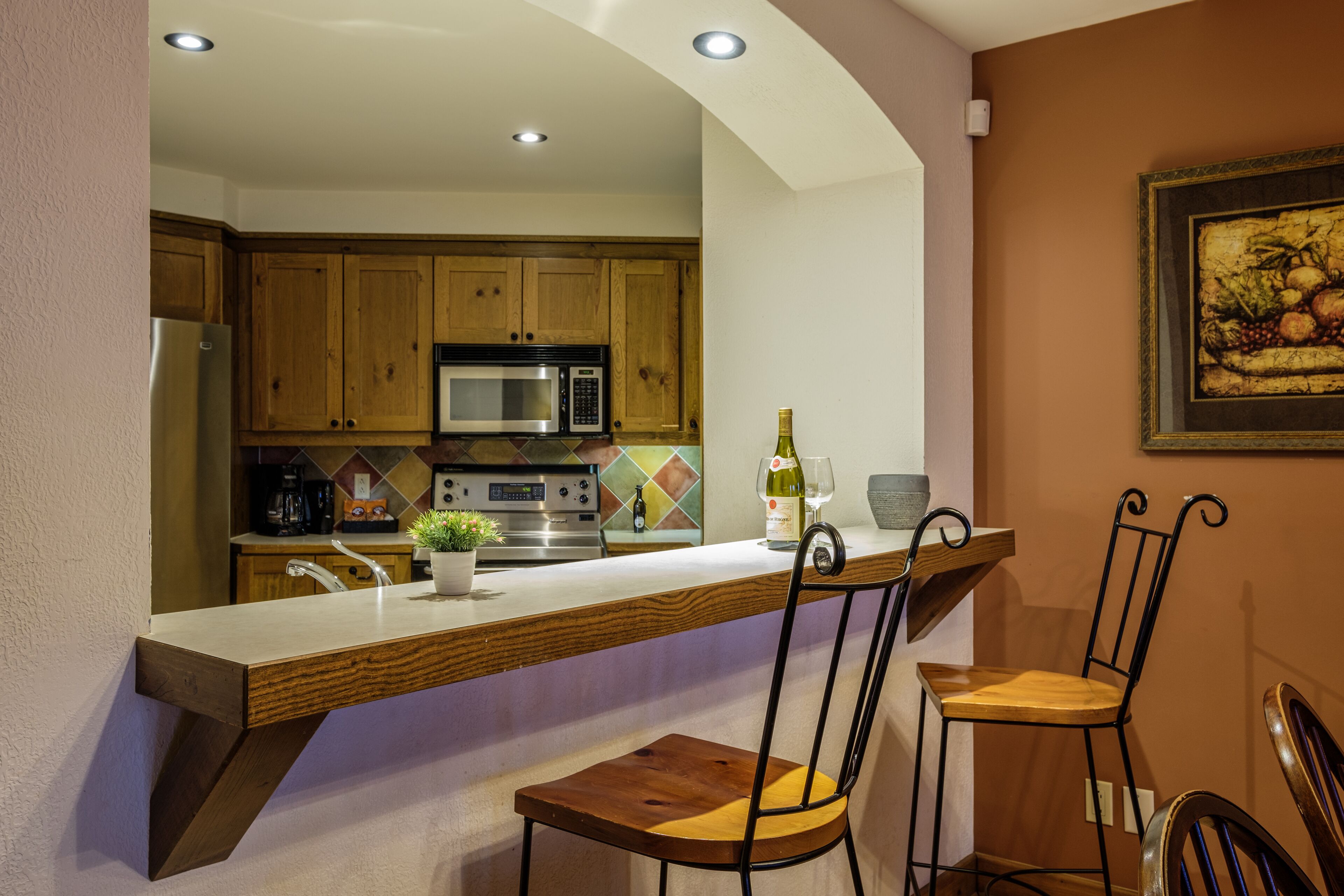 Una cocina con armarios de madera, un refrigerador de acero inoxidable, un microondas y una estufa. Hay una barra con dos sillas de madera.