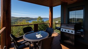 A covered outdoor area with a table and chairs, a grill, and a scenic view of hills and a lake. - L'Equinoxe – RVMT (Mont-Tremblant)