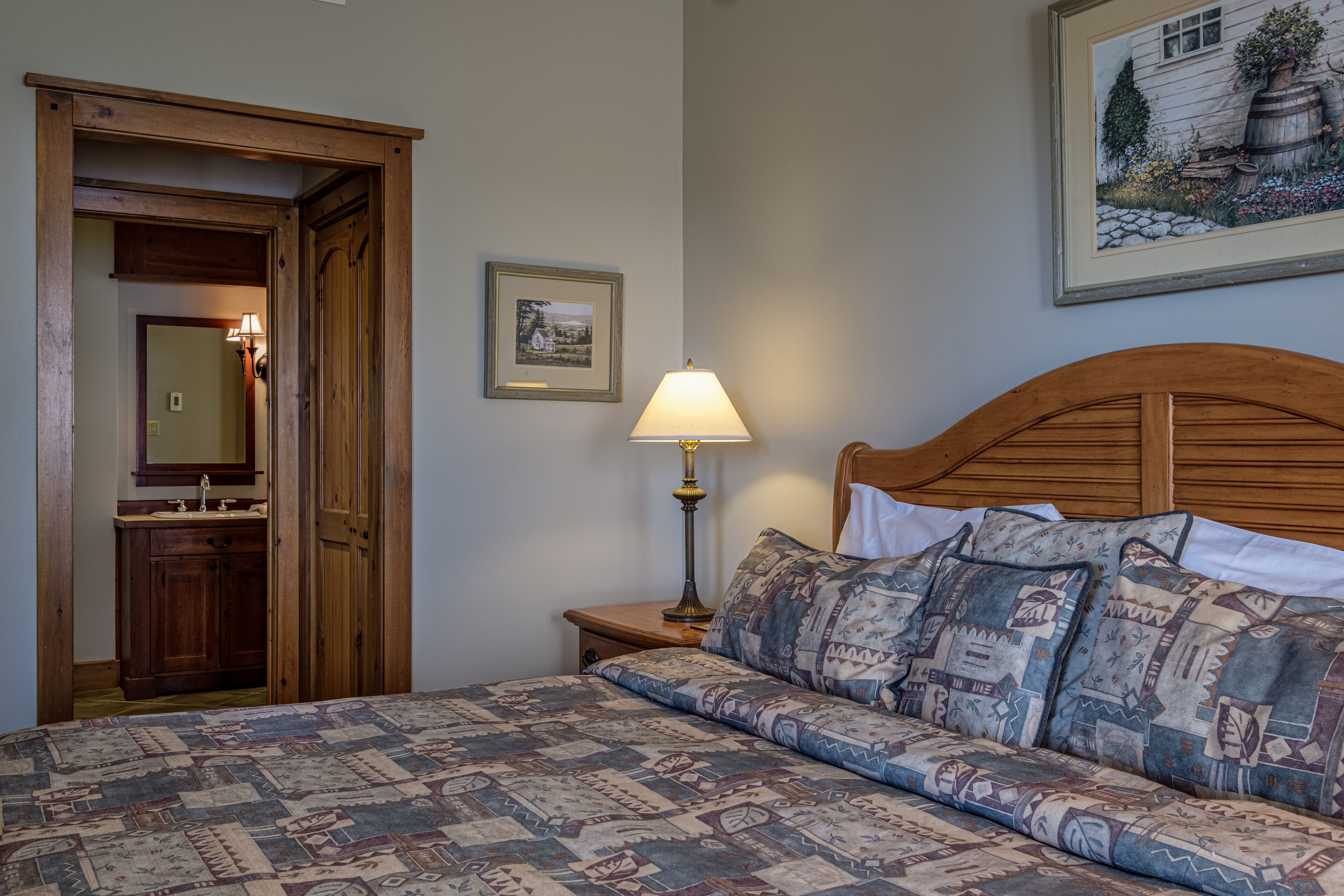 A bedroom with a wooden headboard, a patterned bedspread, a bedside lamp, a framed picture on the wall, and a wooden door leading to a bathroom.