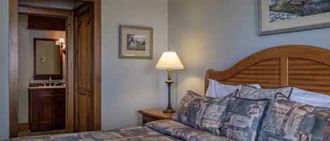 A bedroom with a wooden headboard, a patterned bedspread, a bedside lamp, a framed picture on the wall, and a wooden door leading to a bathroom.