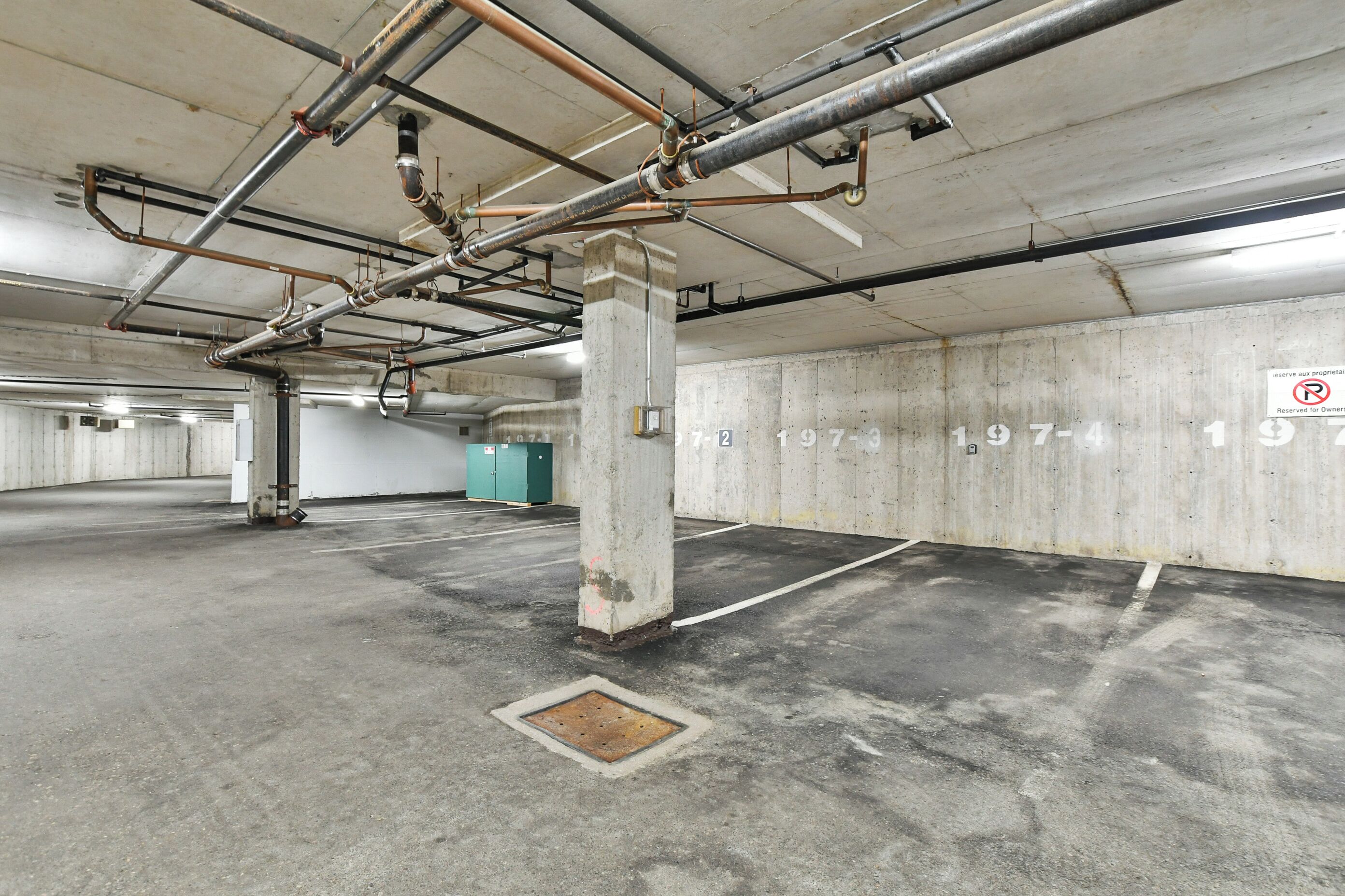 A parking garage with concrete pillars, pipes, and a manhole cover.