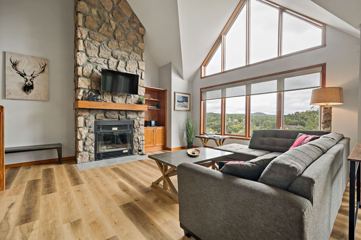 A spacious living room with a stone fireplace, a large window offering a view of the outdoors, a grey sofa, and a wooden coffee table.