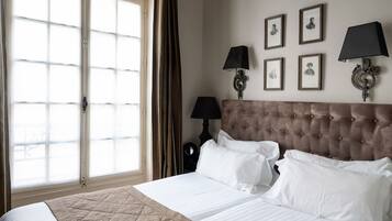 Une chambre d’hôtel avec un grand lit, une tête de lit capitonnée, une fenêtre avec des rideaux et des tableaux encadrés accrochés au mur.