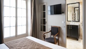 A bedroom with a bed, a chair, a TV, and a bathroom with a sink and mirror. - Hôtel Saint-Louis Marais (Paris)