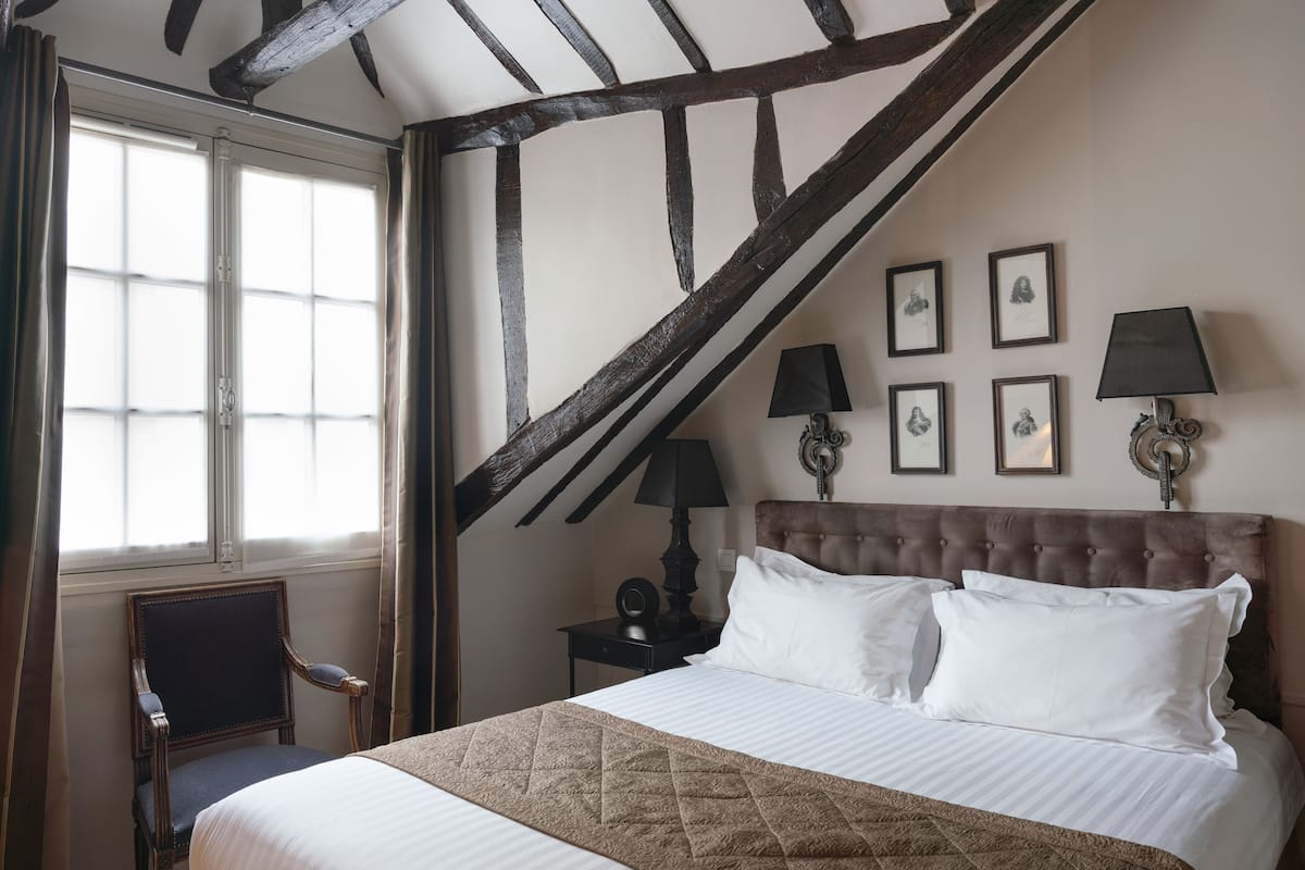 A bedroom with a large bed, a chair, a nightstand with lamps, and framed pictures on the wall.