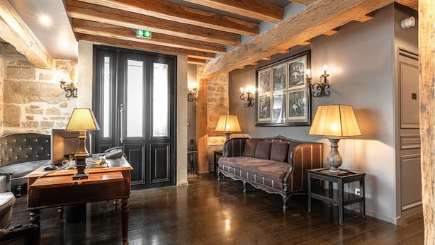 A lobby with wooden beams, a stone wall, a sofa, a table with a lamp, and a door. - Hôtel Saint-Louis Marais (Paris)