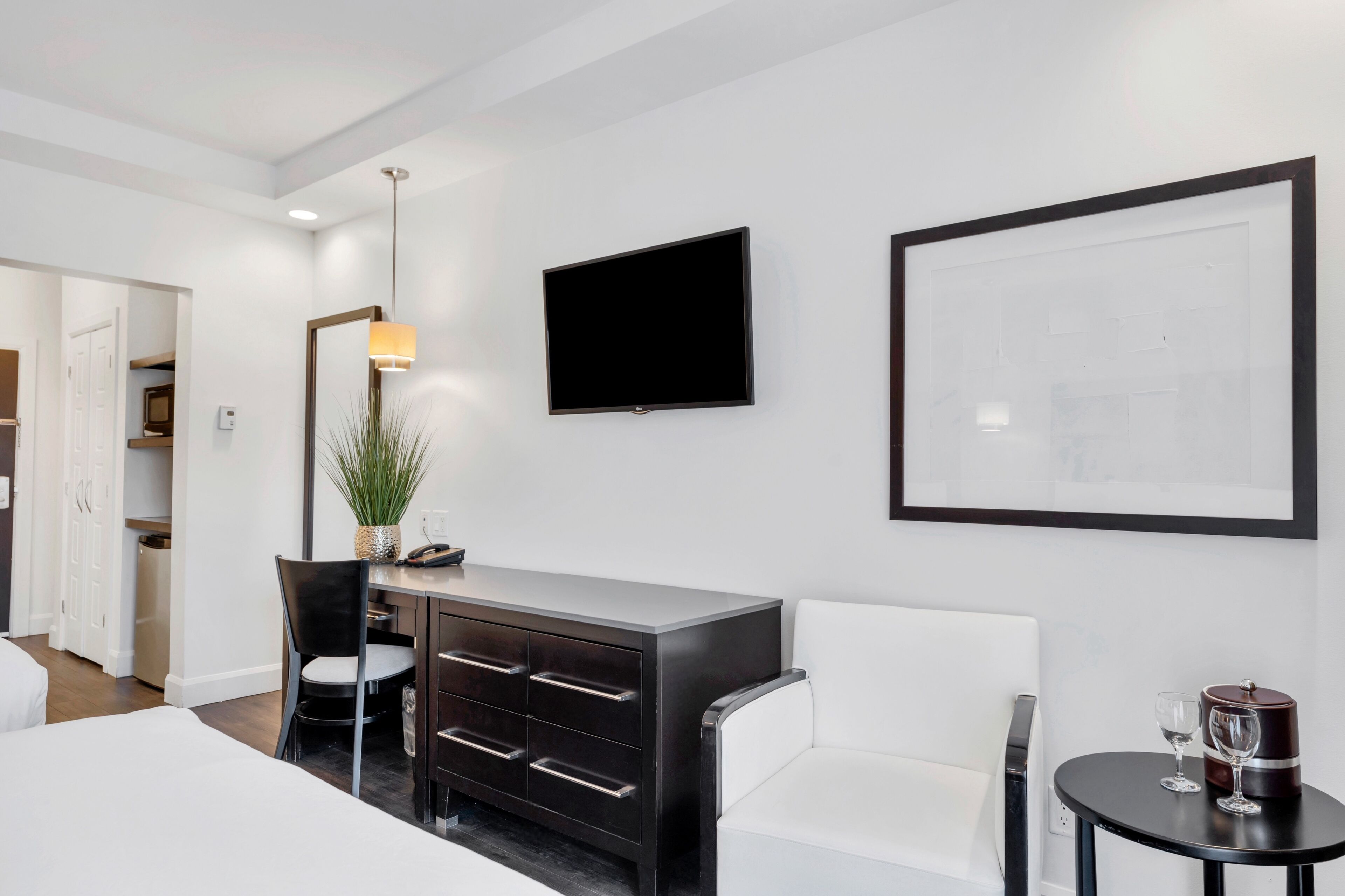 A modern hotel room with a bed, a desk with a chair, a TV, a framed artwork, and a small table with glasses.