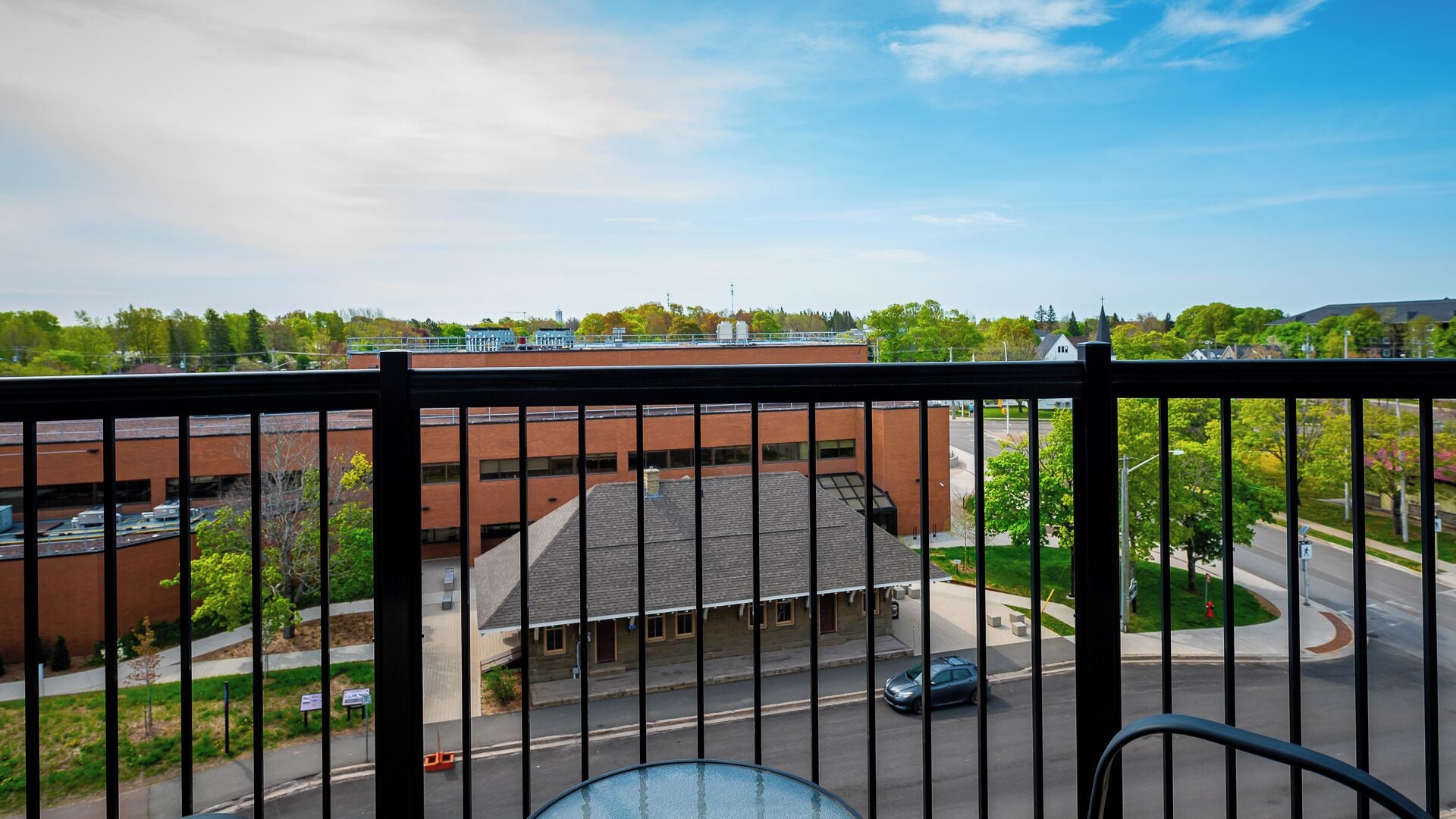 Udsigt fra en balkon med gelænder i metal, der viser en parkeringsplads, en bygning med brun facade og en gade med biler og grønt.