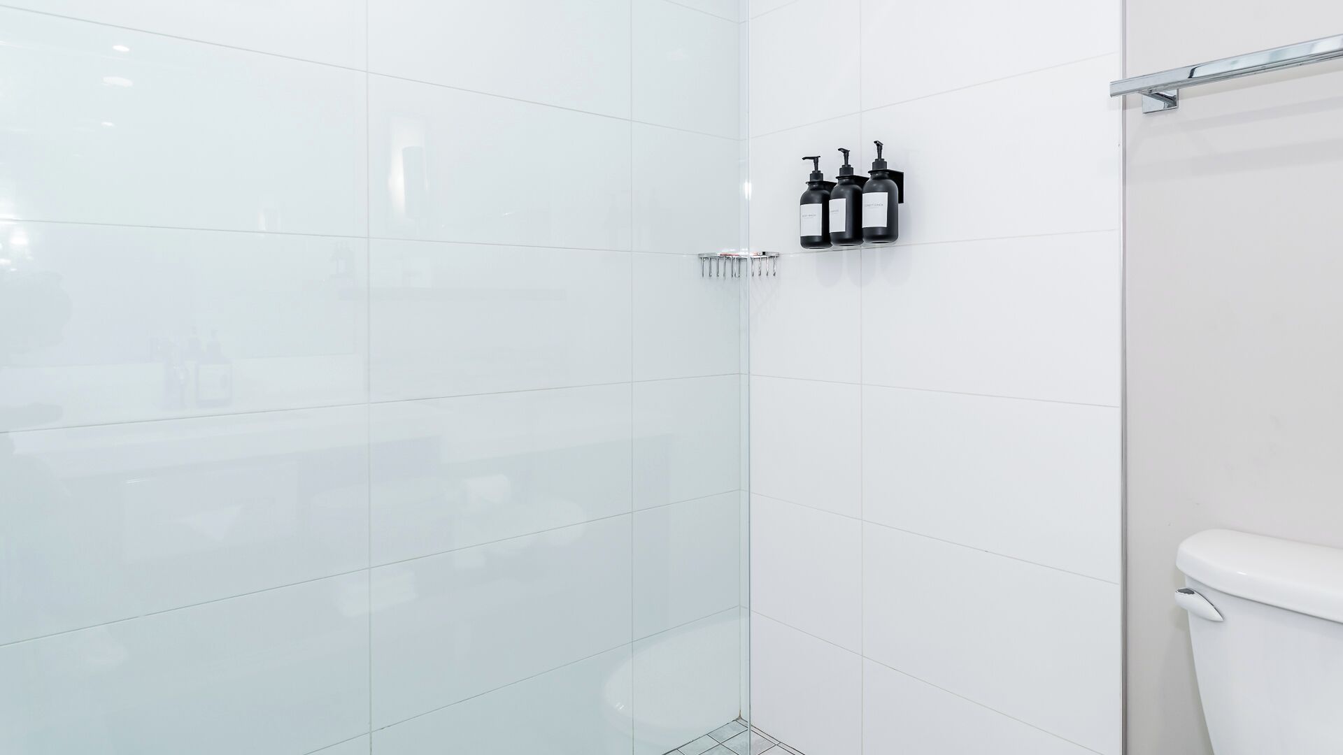 A modern bathroom with white tiles, a glass shower enclosure, and black toiletries on a wall-mounted rack.